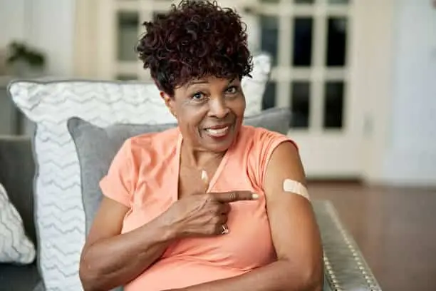 Waist-up view of casually dressed woman sitting in family home and smiling at camera as she gestures with pride at adhesive bandage.